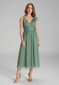 Grünes, ärmelloses Kleid mit tiefem V-Ausschnitt und Stoffblumenakzent an der Schulter. Fließender Rock mit tailliertem Bund. Trägt goldene High Heels.