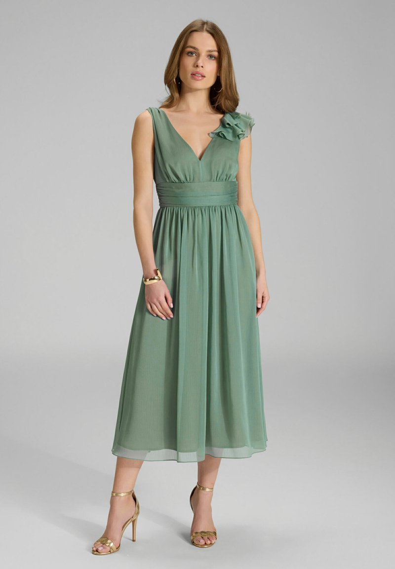 Grünes, ärmelloses Kleid mit tiefem V-Ausschnitt und Stoffblumenakzent an der Schulter. Fließender Rock mit tailliertem Bund. Trägt goldene High Heels.