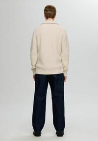 Suéter de punto color crema con textura acanalada, con un cuello vuelto. Combinado con unos jeans de pierna ancha de color azul oscuro y zapatos negros.