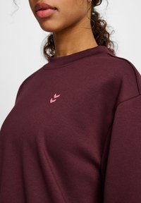 Bordeaux sweatshirt gemaakt van zacht materiaal, met een hoge halslijn en een klein roze logo op de borst. Gladde textuur met een relaxte pasvorm.
