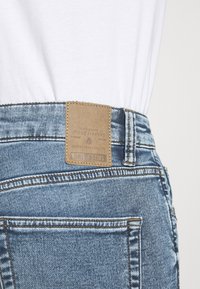 Jeans de mezclilla azul claro con un parche de cuero marrón en la cintura, que presentan detalles de costura y diseño en el bolsillo trasero.
