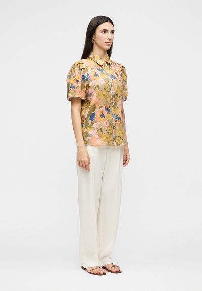 Γυναίκα όρθια, φορούν κοντομάνικο φλοράλ μπλουζάκι με φουσκωτούς ώμους, λευκό παντελόνι με φαρδιά ποδόγυρα, και καφέ σανδάλια.