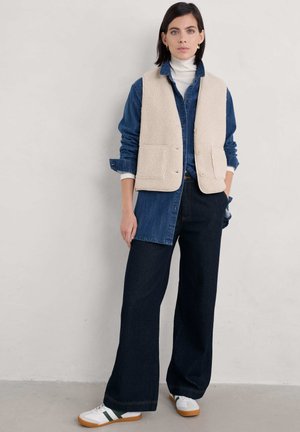 Beige fleece vest over a denim shirt, paired with wide-leg dark jeans and white sneakers with green accents; minimalistic design, casual look.