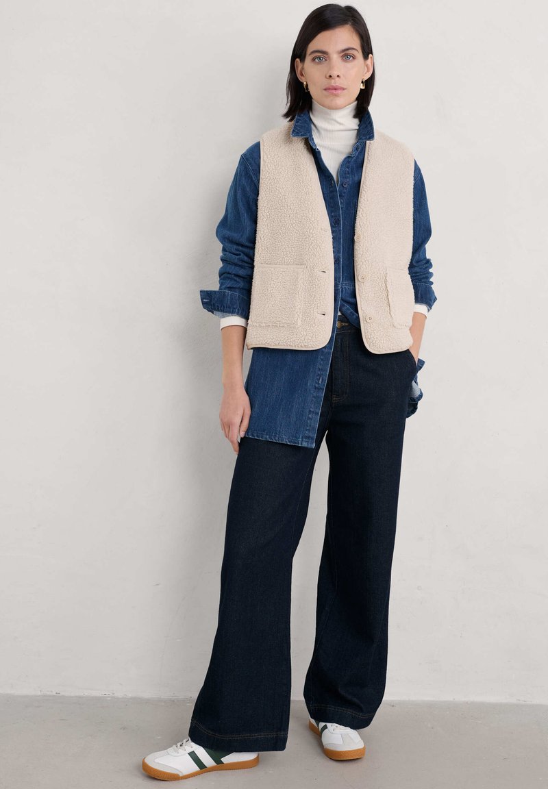 Bežna flis brezrokavnica čez jeans srajco, skupaj s širokimi temnimi kavbojkami in belimi supergami z zelenimi detajli; minimalističen slog, sproščen videz.