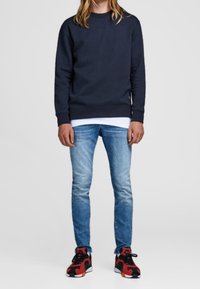 Sweatshirt bleu marine, jeans ajustés bleu clair et baskets noir-rouge-orange. Le sweatshirt a des poignets et un col côtelés.