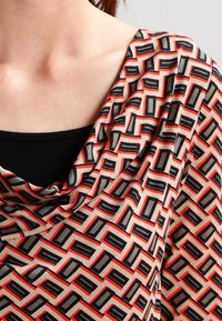 Wzorzysty top z dekoltem z drapowaniem; ozdobiony geometrycznym wzorem w kolorach czerwonym, czarnym i beżowym, z gładką fakturą materiału i subtelnym połyskiem.