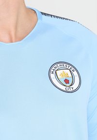 Helles blaues Sporttrikot mit Rundhalsausschnitt und kurzen Ärmeln, auf dem die Manchester-City-Logostickerei auf der Brust prangt.