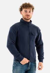 Felpa blu navy con zip, colletto alto, tasca anteriore, polsini e orlo a costine; realizzata in tessuto morbido con una superficie liscia.