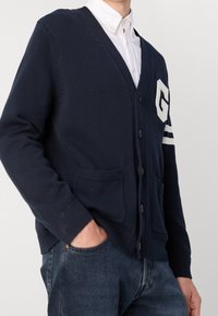 Marinblå stickad cardigan med v-ringning, knäppning, två fickor och vit "G"-emblem med ränder på ärmarna. Material i bomullsblandning.