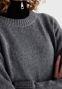 Szary wełniany sweter z prążkowanym dekoltem i mankietami, z czarnym zamkiem b�łyskawicznym i stójką. Tekstura jest miękka i lekko puszysta.