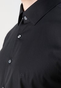 Gros plan sur une personne portant une chemise noire à boutons avec un col pointu et un petit logo brodé "ck" sur la poitrine.