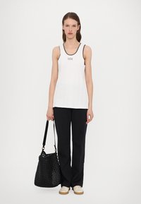 White sleeveless tank top with dark trim, featuring "MM" embroidery, paired with black trousers and a woven black bucket bag. Casual footwear.