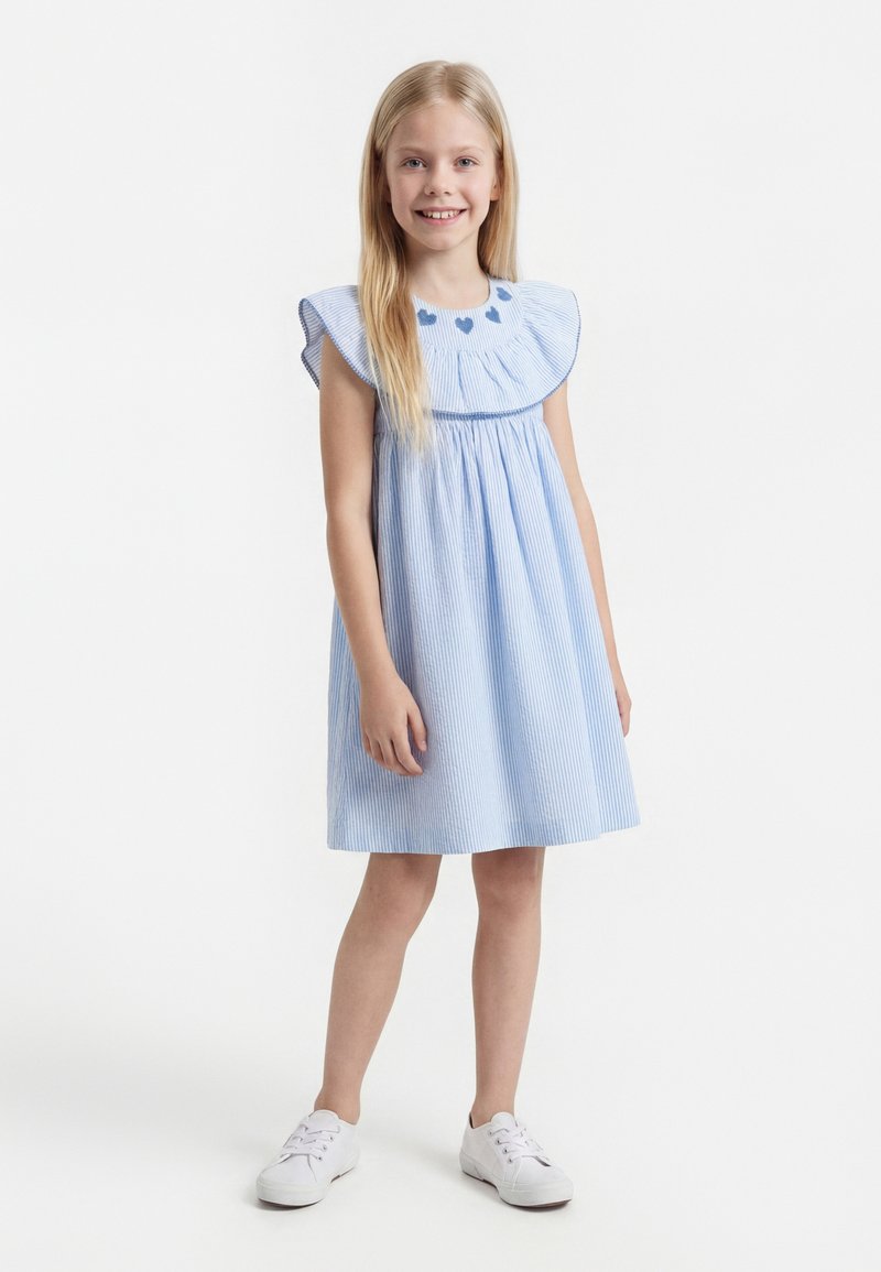 Νεαρή κοπέλα στέκεται, φορώντας ένα ανοιχτό μπλε ριγέ φόρεμα με φουσκωτά μανίκια και λεπτομέρειες καρδιάς στο ντεκολτέ, συνδυασμένο με λευκά αθλητικά παπούτσια.