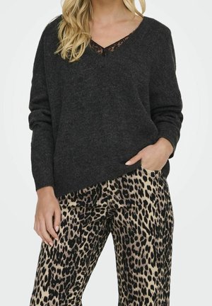 Frau trägt dunkelgrauen V-Ausschnitt-Pullover mit Spitzenbesatz und Leopardenmuster-Hose, steht mit einer Hand in der Tasche.
