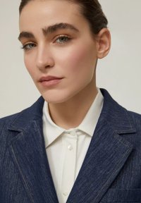 Blazer in denim blu scuro con cuciture arancioni a contrasto, indossato sopra una camicia bianca con colletto, caratterizzato da texture morbide e un design strutturato.