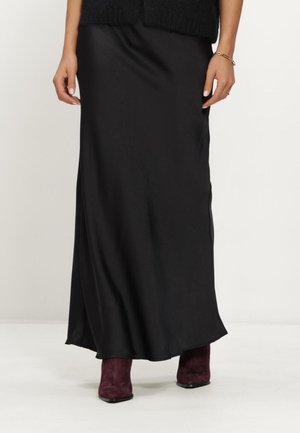 Woman wearing a long black satin skirt paired with dark purple suede ankle boots standing on a white background.