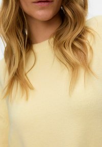 Pull tricoté jaune clair avec un col côtelé, une texture douce et une coupe décontractée, partiellement visible avec de longs cheveux ondulés en arrière-plan.