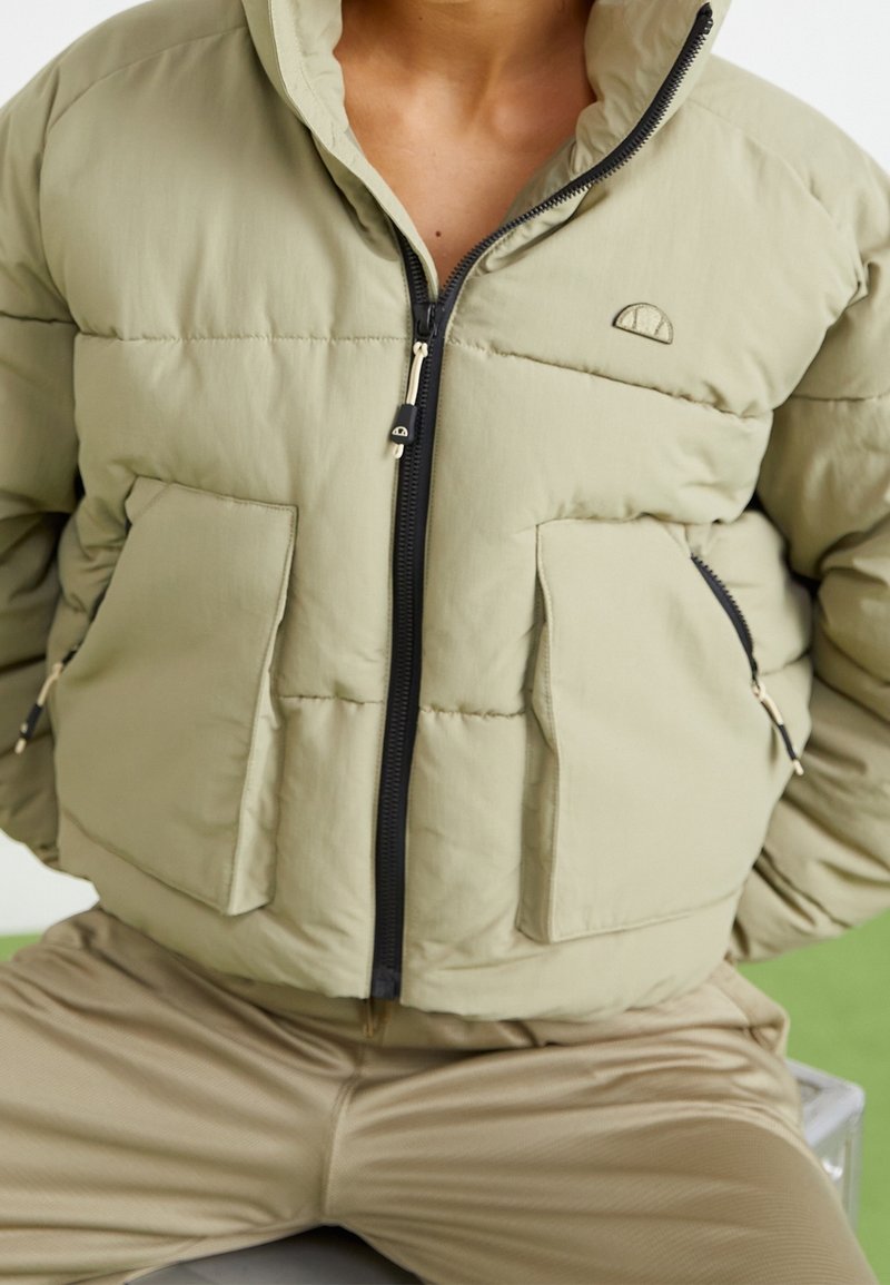 Puffer jacket in light olive green, featuring a zip front, large pockets, and an adjustable hood with a subtle logo detail on the chest.