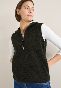 Fleece-Weste in Dunkelgrün mit Reißverschluss am Kragen, mit strukturierter Oberfläche und seitlichen Taschen, kombiniert mit einem weißen Langarmshirt.