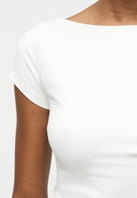 Κοντινό πλάνο ενός ατόμου που φοράει ένα απλό λευκό στενό κοντομάνικο μπλουζάκι με γυρισμένα μανίκια.