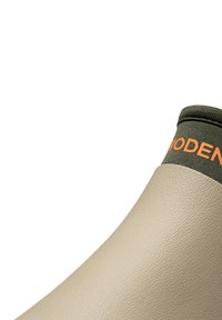 Gumowa boot z zakrzywioną beżową powierzchnią zewnętrzną i teksturowanym wykończeniem. Górna część ma oliwkową mankietę z pomarańczowym napisem marki.