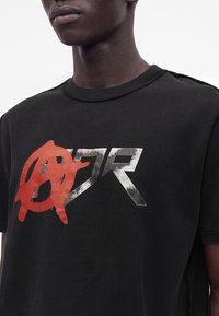 Κοντινό πλάνο ενός ατόμου που φοράει μαύρο T-shirt με ένα κόκκινο σύμβολο αναρχίας που καλύπτει ασημένια φθαρμένα γράμματα "ZR" στο μπροστινό μέρος.
