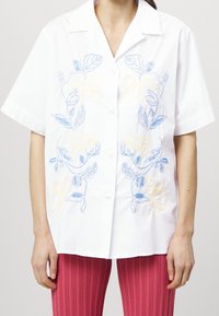 Camicia bianca a maniche corte con ricami floreali blu e gialli, chiusura con bottoni, vestibilità rilassata e texture in cotone morbido.