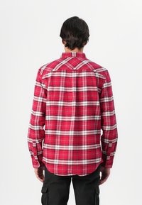 Camisa de cuadros rojos con detalles en blanco, cuenta con mangas largas, un dobladillo curvado y dos piezas en la parte posterior. La tela parece ser tejida y con textura.