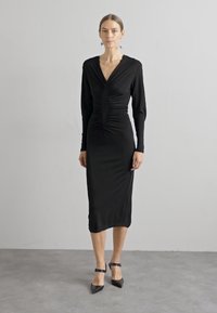 Μαύρο φόρεμα μακρύ μανίκι με βαθύ V-λαιμόκοψη, συγκεντρωμένες λεπτομέρειες στη μέση, slim fit και μήκος πάνω από το γόνατο. Συνδυασμένο με μαύρες στενές γόβες.