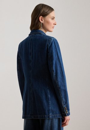 Femme portant un blazer en denim bleu foncé avec des boutons dorés sur le revers, vue de profil sur un fond uni clair.