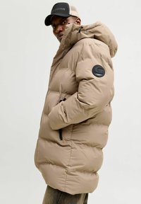 Mies, joka käyttää beigeä toppatakki hupulla ja mustaa sekä beigen väristä lippalakkia, seisoo sivuprofiilissa kädet taskuissa.