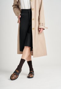 Manteau beige avec des boutons dorés, associé à une jupe noire et des chaussures à motifs marron. Chaussettes marron foncé visibles. Fond simple.