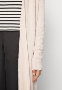 Jasnoróżowy sweter robiony na drutach z długimi rękawami i ściągaczami noszony na czarno-białą, paskowaną bluzkę, zestawiony z ciemnymi spodniami.