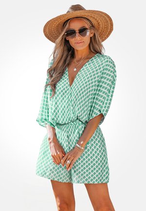 Mujer con un mono verde y blanco de estampado, sombrero de paja, gafas de sol y joyas doradas sobre un fondo blanco liso.