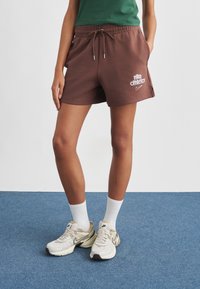 Καφέ αθλητικό σορτς Nike με ελαστική μέση και κορδόνι. Συνδυασμένο με λευκές κάλτσες μέσης και μπεζ αθλητικά παπούτσια Nike σε μπλε φόντο.