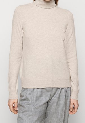 Personne portant un pull à col roulé beige clair et un pantalon gris ample debout devant un fond uni.