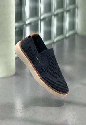 Slip-on shoes in dark navy mesh material with textured accents, cream rubber sole, and tan detail at the heel. Lightweight design.