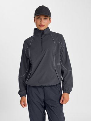 Vrouw draagt een donkergrijze jas met een kwartritssluiting, bijpassende broek met reflecterende bies en een zwarte baseballpet tegen een effen achtergrond.