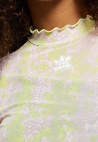 Nahaufnahme einer Person, die ein hellgrünes und pink gemustertes Adidas-Oberteil mit Rüschensaum und sichtbar aufgenähtem Adidas-Logo auf der Brust trägt.