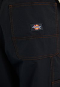 Pantalons de travail noirs en tissu avec des surpiqûres orange, comprenant une poche latérale et un patch logo Dickies visible.