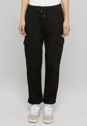 Person trägt schwarze Cargo-Hose mit Kordelzugbund und graue Adidas-Sneaker, steht vor hellem, einfarbigem Hintergrund.