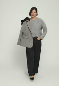 Maglione grigio a coste, blazer grigio con tasca e pantaloni a gamba larga neri e testurizzati. La modella indossa scarpe nere e tiene il blazer su una spalla.