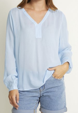 Kobieta ubrana w jasnoniebieską bluzkę z długim rękawem i dekoltem w serek, swobodnie włożoną do podwiniętych szortów dżinsowych, z jedną ręką w kieszeni.