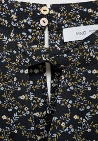 Zwarte bloemenblouse met kleine witte, blauwe en gele bloemen, voorzien van een sleutelgatopening en twee knoopsluitingen bij de hals.