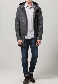 Parka imperméable gris foncé avec capuche, fermetures à pression, cordons réglables et poches latérales, portée sur une chemise gris clair et un jean bleu.