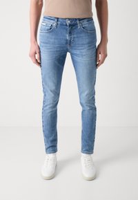 Jeans de mezclilla de color azul claro con un diseño ajustado, que cuentan con un cierre de botón, cinco bolsillos y un desgaste sutil en los muslos. Combinados con zapatillas blancas.