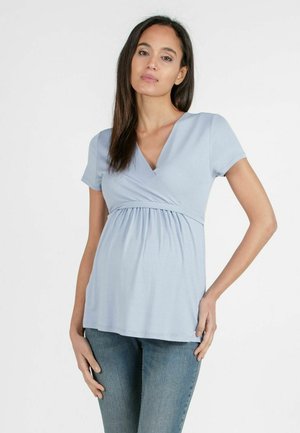 Gravid kvinde med langt mørkt hår iført en lyseblå kortærmet slå-om graviditetstop og blå jeans, stående med hånd på hoften.