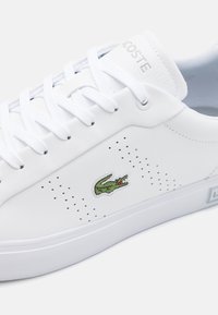 Baskets blanches Lacoste avec lacets blancs, évents latéraux perforés et logo crocodile vert sur le panneau latéral.
