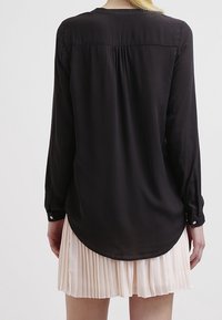 Blusa nera a maniche lunghe con una texture morbida e liscia e orlo arrotondato. Presenta un colletto arricciato sul retro e polsini con bottoni. Abbinata a una gonna plissettata.