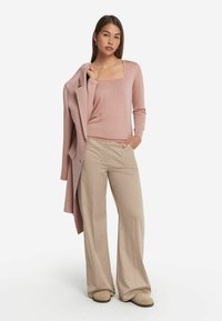 Mjukt rosa långärmad topp med fyrkantig halsringning, kombinerad med beige vida byxor och en matchande kappa som hänger över ena armen.